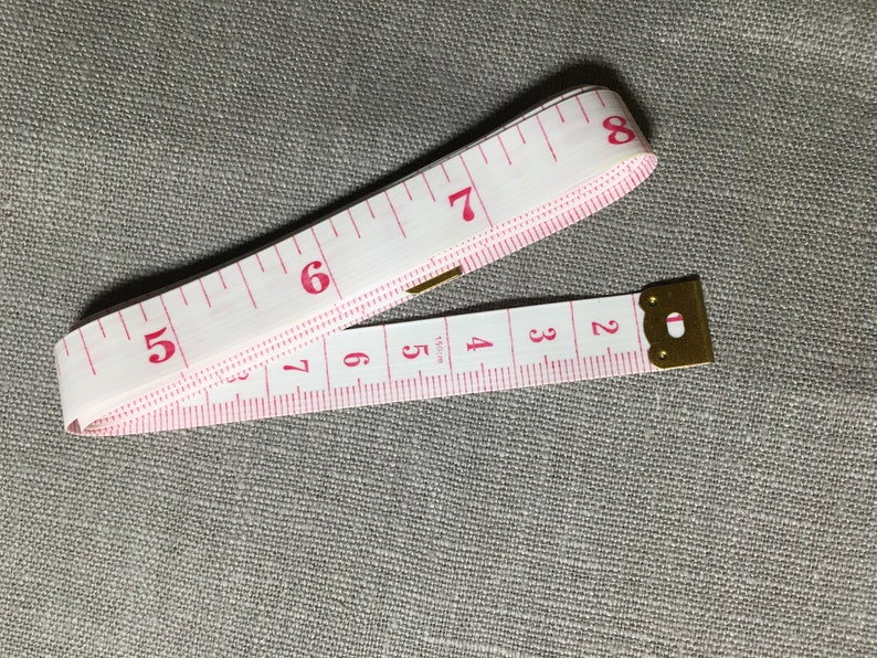 May include: A white measuring tape with pink markings and a gold metal end. The tape is extended to show numbers 1 through 8.