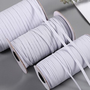 May include: Three spools of white elastic cord, wound on cardboard spools with black labels. The elastic cord is a popular material for crafting and sewing projects.