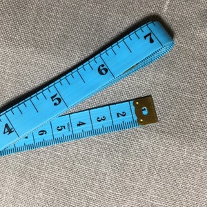 May include: A blue measuring tape with black numbers from 2 to 7. The tape is folded in half and the end is secured with a gold metal clasp.