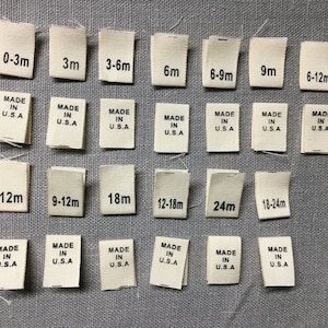 May include: A selection of off-white fabric labels with black text denoting baby clothing sizes, such as 0-3m, 3m, 3-6m, 6m, 6-9m, 9m, 6-12m, 12m, 9-12m, 18m, 12-18m, 24m, and 18-24m. Each label is marked "MADE IN U.S.A."