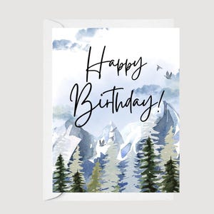 May include: A white greeting card with a watercolor illustration of a mountain range and evergreen trees. The card features the words "Happy Birthday!" in black script. The background has a blue and white sky.