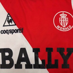 May include: A white and red soccer jersey with a black "BALLY" logo across the front. The jersey features the Le Coq Sportif logo on the left side and the AS Monaco logo on the right side.