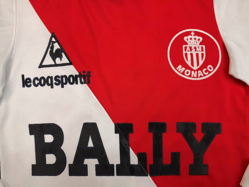 May include: A white and red soccer jersey with a black "BALLY" logo across the front. The jersey features the Le Coq Sportif logo on the left side and the AS Monaco logo on the right side.