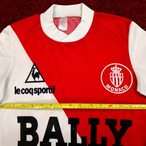 May include: A white and red long-sleeved soccer jersey with the text "le coq sportif" and a rooster logo on the left side. The right side of the jersey has the text "AS Monaco" and a crest with a crown. The jersey has a black "BALLY" logo across the bottom.