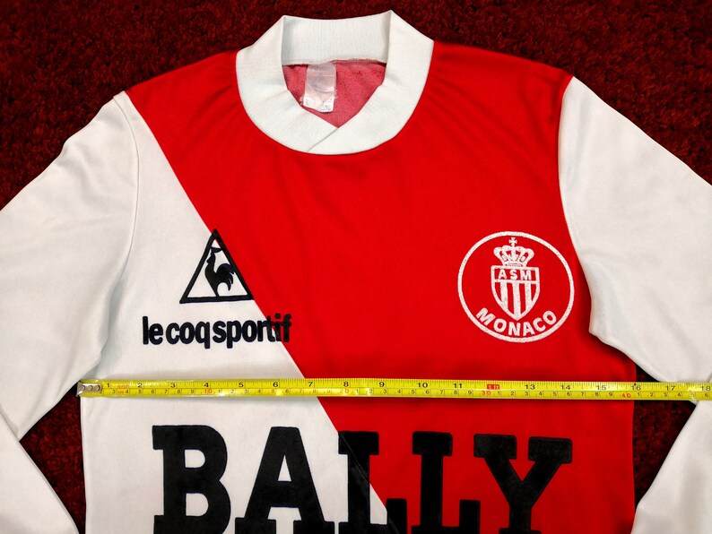 May include: A white and red long-sleeved soccer jersey with the text "le coq sportif" and a rooster logo on the left side. The right side of the jersey has the text "AS Monaco" and a crest with a crown. The jersey has a black "BALLY" logo across the bottom.