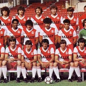 May include: A group of 20 men wearing red and white striped soccer jerseys with the word "BALLY" printed on them. The men are standing in two rows, with the front row sitting on a bench. The men are all smiling and looking at the camera.