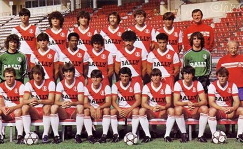 May include: A group of 20 men wearing red and white striped soccer jerseys with the word "BALLY" printed on them. The men are standing in two rows, with the front row sitting on a bench. The men are all smiling and looking at the camera.