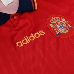 May include: Red soccer jersey with a gold adidas logo and the Spanish national team crest. The jersey has a blue collar with a yellow stripe.