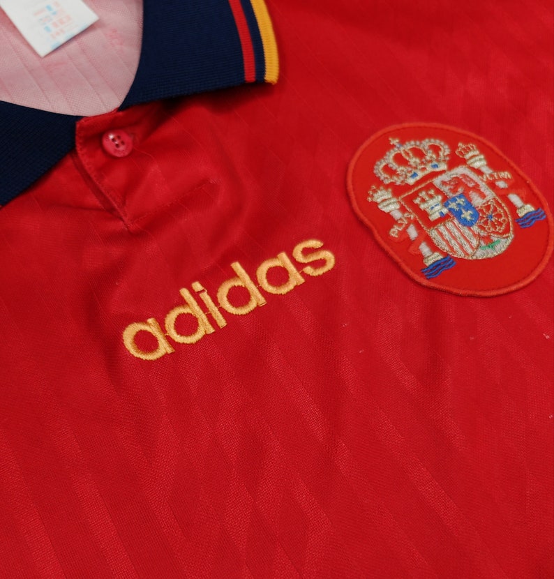May include: Red soccer jersey with a gold adidas logo and the Spanish national team crest. The jersey has a blue collar with a yellow stripe.