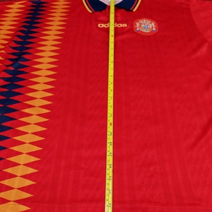 May include: Red soccer jersey with a diamond pattern on the left side, the Adidas logo, and the Spanish national team crest. The jersey is being measured with a tape measure, showing a length of 31 inches.