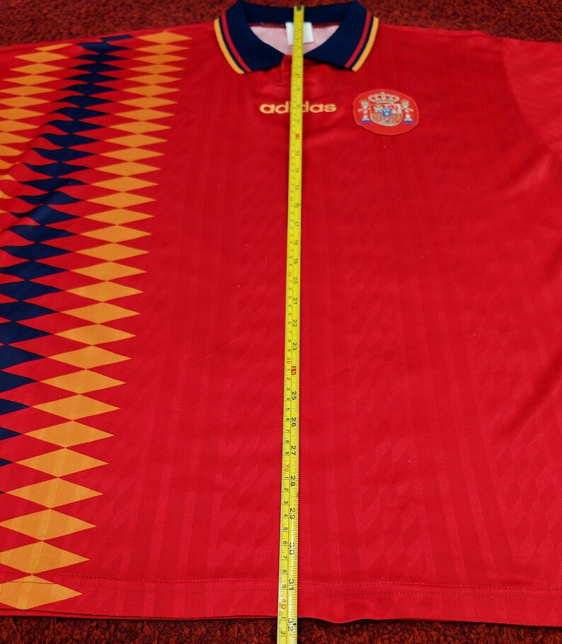 May include: Red soccer jersey with a diamond pattern on the left side, the Adidas logo, and the Spanish national team crest. The jersey is being measured with a tape measure, showing a length of 31 inches.