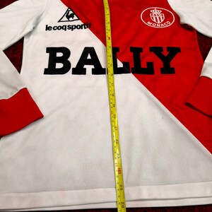 May include: White and red long-sleeve soccer jersey with a black rooster logo, the text "le coq sportif", and the text "BALLY" in large black letters. The jersey also features a red crest with the text "ASM MONACO".