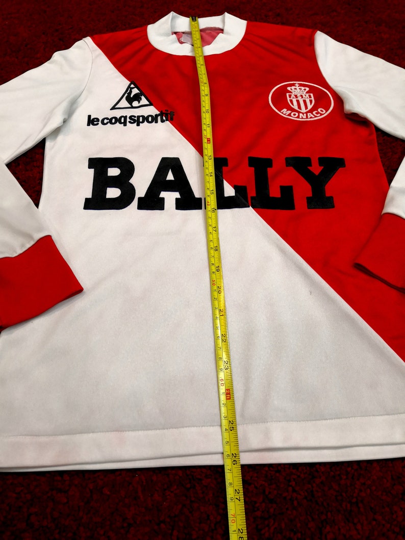 May include: White and red long-sleeve soccer jersey with a black rooster logo, the text "le coq sportif", and the text "BALLY" in large black letters. The jersey also features a red crest with the text "ASM MONACO".