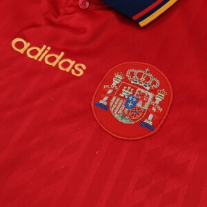May include: Red soccer jersey with a yellow adidas logo and a red embroidered crest of the Spanish national team.