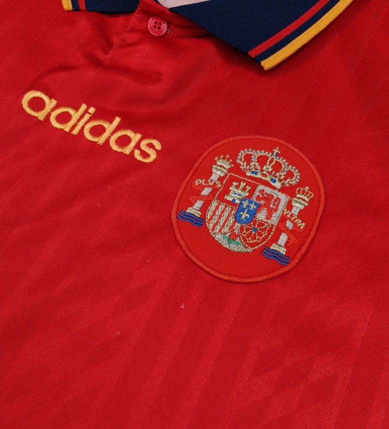 May include: Red soccer jersey with a yellow adidas logo and a red embroidered crest of the Spanish national team.