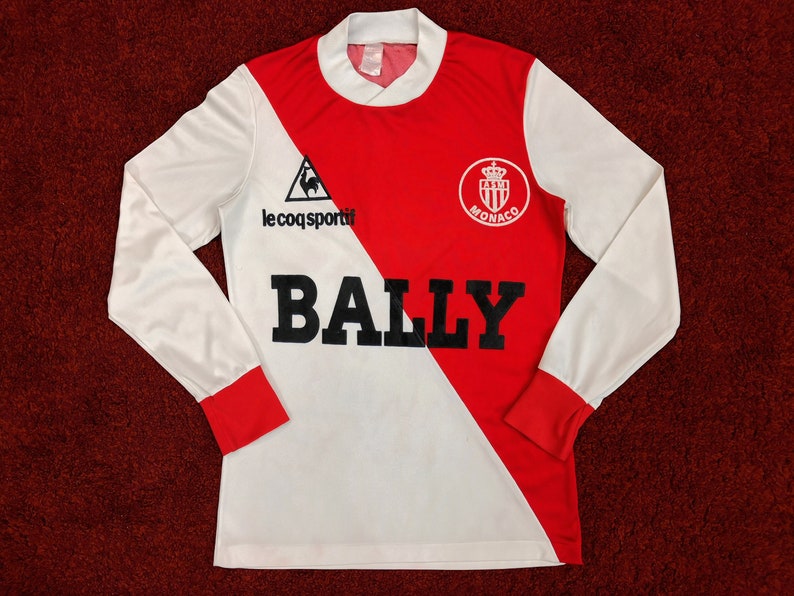 May include: White and red long-sleeve soccer jersey with a diagonal split. The jersey features the Le Coq Sportif logo, the AS Monaco crest, and the word "BALLY" in large black letters.