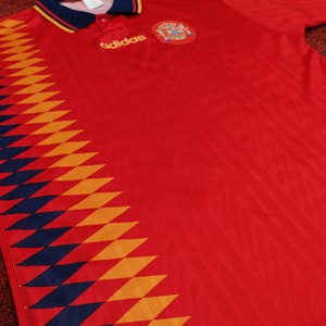 May include: Red soccer jersey with a blue and yellow diamond pattern on the left side. The jersey has the Adidas logo and the Spanish national team crest on the chest.