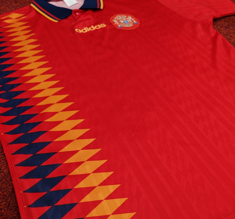 May include: Red soccer jersey with a blue and yellow diamond pattern on the left side. The jersey has the Adidas logo and the Spanish national team crest on the chest.