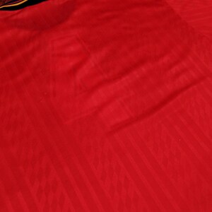 May include: Red soccer jersey with a textured pattern. The jersey has a yellow and blue stripe across the shoulders.