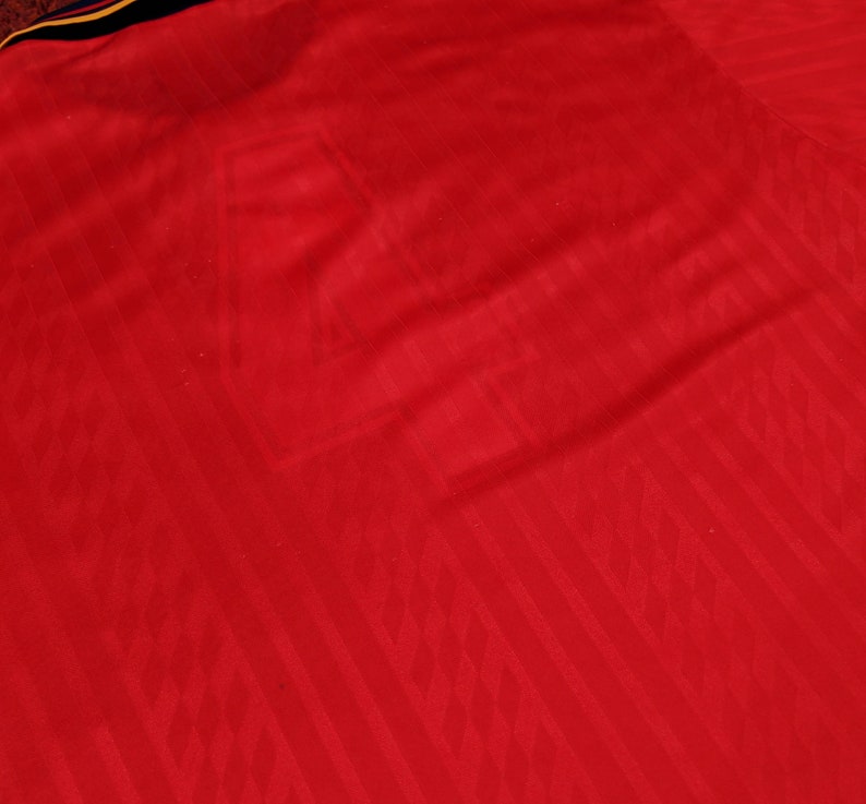 May include: Red soccer jersey with a textured pattern. The jersey has a yellow and blue stripe across the shoulders.