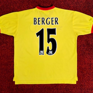 May include: A yellow soccer jersey with the name "BERGER" and the number "15" in black. The jersey has red and black trim on the collar and sleeves. The background is a dark red, textured surface.