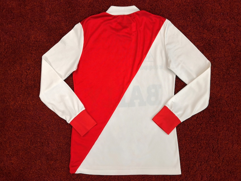May include: A white and red long-sleeved jersey with a diagonal red stripe. The jersey has a white collar and red trim on the sleeves. The number "8" is printed on the back of the jersey.