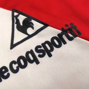 May include: A white and red athletic jersey with a black rooster logo and the text "le coq sportif" in black.