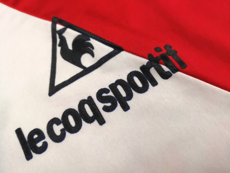 May include: A white and red athletic jersey with a black rooster logo and the text "le coq sportif" in black.