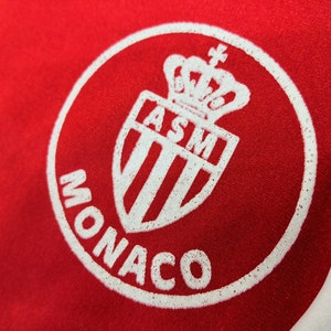 May include: White and red circular logo with the text "MONACO" and a white shield with a crown and the letters "ASM" inside.