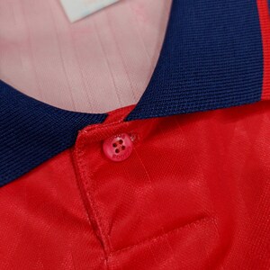 May include: Close-up of a red soccer jersey with a navy blue collar and yellow trim. The collar has a button closure with a white button that says "adidas" in black.