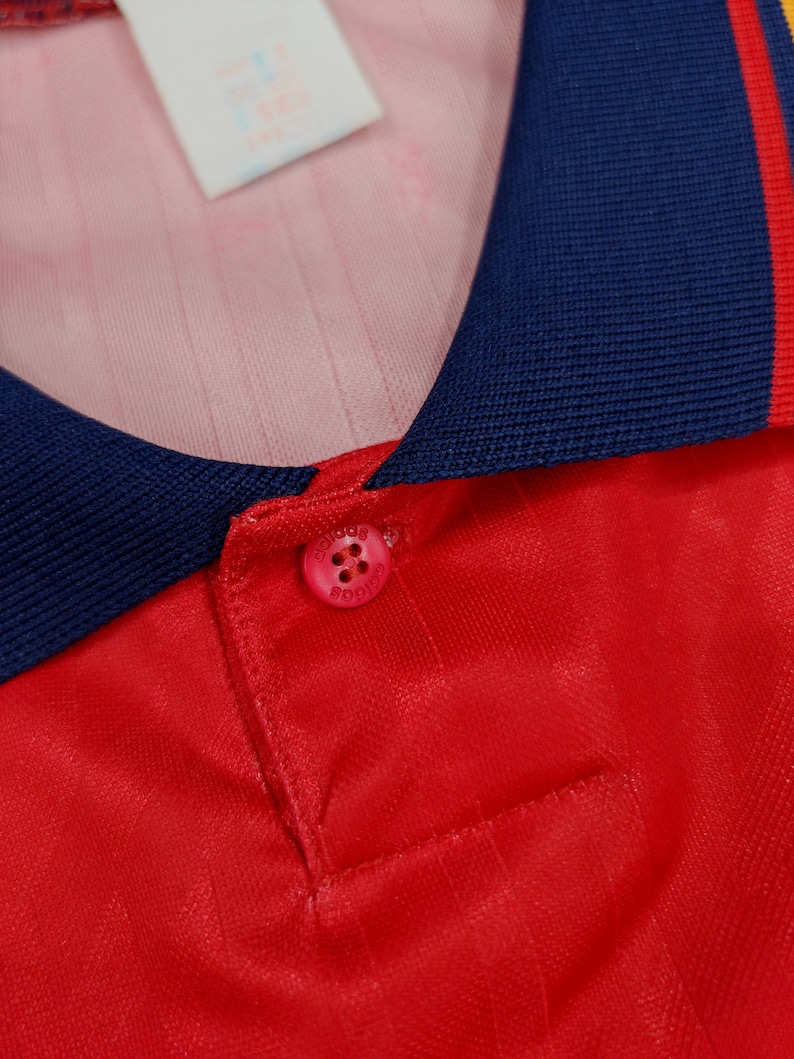 May include: Close-up of a red soccer jersey with a navy blue collar and yellow trim. The collar has a button closure with a white button that says "adidas" in black.