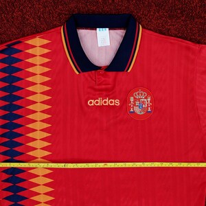 May include: Red soccer jersey with a blue and yellow diamond pattern on the left side. The jersey has a black collar with a red stripe and the Adidas logo on the chest. The Spanish national team crest is on the right side of the chest.