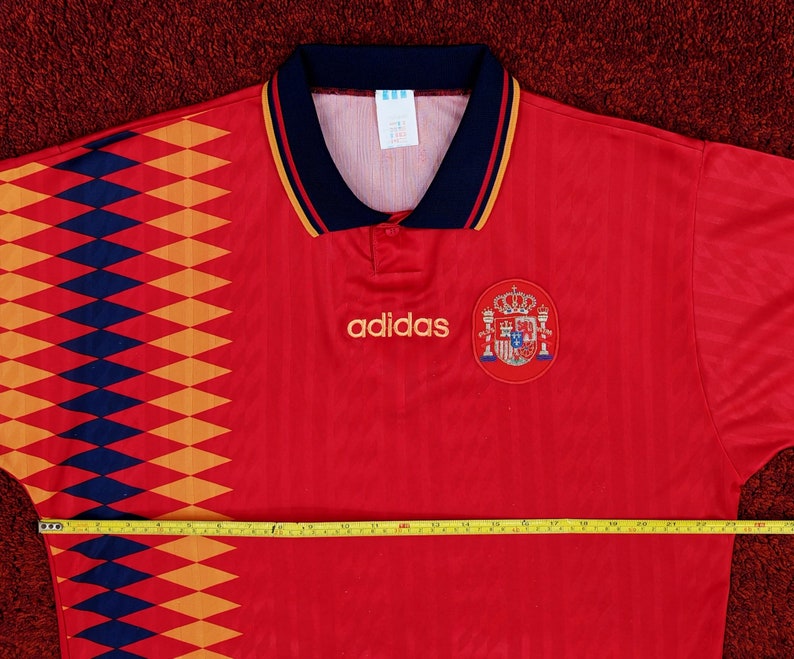May include: Red soccer jersey with a blue and yellow diamond pattern on the left side. The jersey has a black collar with a red stripe and the Adidas logo on the chest. The Spanish national team crest is on the right side of the chest.