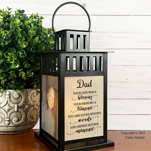 May include: Black metal lantern with a photo and text that reads "Dad, Your life was a blessing, Your memory a treasure, You are loved beyond words, and missed beyond measure." The lantern has a handle and sits on a base.