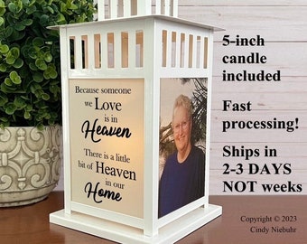 May include: White metal lantern with a handle, featuring a photo and the text "Because someone we Love is in Heaven...in our Home." Includes a 5-inch candle. The lantern is on a wooden surface.