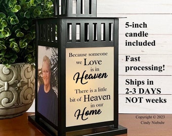 May include: Black metal lantern with a handle, featuring a photo and the text "Because someone we Love is in Heaven." Includes a 5-inch candle. The lantern is displayed near a green plant in a decorative pot.