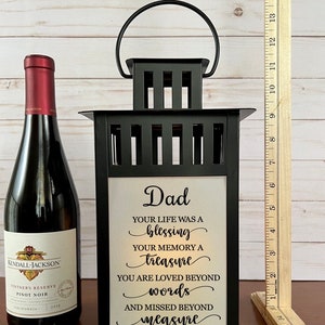 May include: A black metal lantern with a handle, featuring a white panel with the text "Dad" and a heartfelt message. A bottle of Kendall-Jackson Pinot Noir wine is to the left. A wooden ruler is to the right.