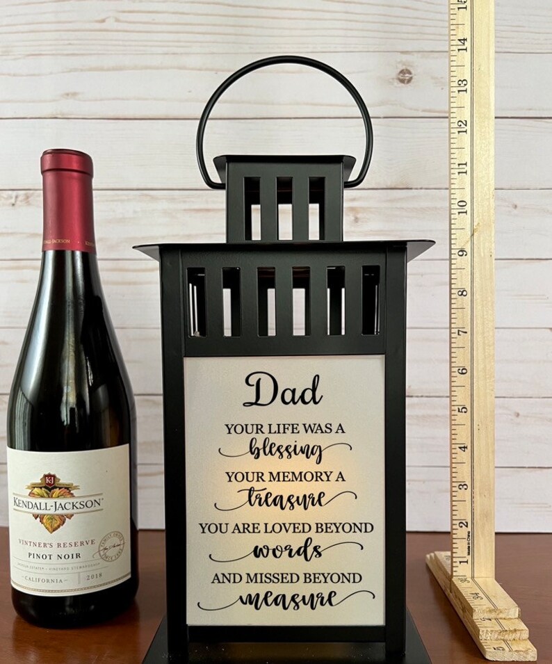 May include: A black metal lantern with a handle, featuring a white panel with the text "Dad" and a heartfelt message. A bottle of Kendall-Jackson Pinot Noir wine is to the left. A wooden ruler is to the right.
