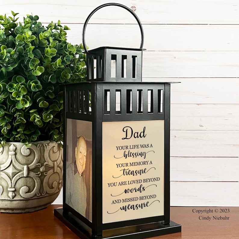 May include: Black metal lantern with a photo and text that reads "Dad, Your life was a blessing, Your memory a treasure, You are loved beyond words, and missed beyond measure." The lantern has a handle and sits on a base.