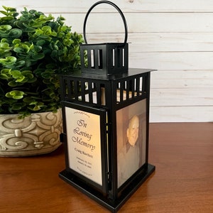 May include: Black metal memorial lantern with a handle, featuring a photo and text that reads "In Loving Memory Craig Bartlett". The lantern has a square top and base, with a decorative design.