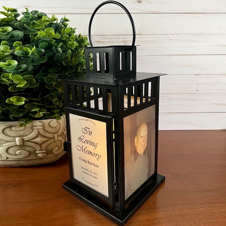 May include: Black metal memorial lantern with a handle, featuring a photo and text that reads "In Loving Memory Craig Bartlett". The lantern has a square top and base, with a decorative design.