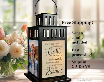 May include: A black metal lantern featuring a top handle and a square frame. One side displays a photograph of a person with a beard, while the adjacent side features the text: There are some who bring a Light so great to the world that even after they have gone the light Remains.