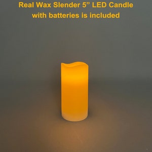 May include: A slender, real wax LED candle, approximately 5 inches tall, with a softly glowing amber light. The candle has a slightly wavy top edge and a frosted base. Text on the image reads: "Real Wax Slender 5" LED Candle with batteries is included."