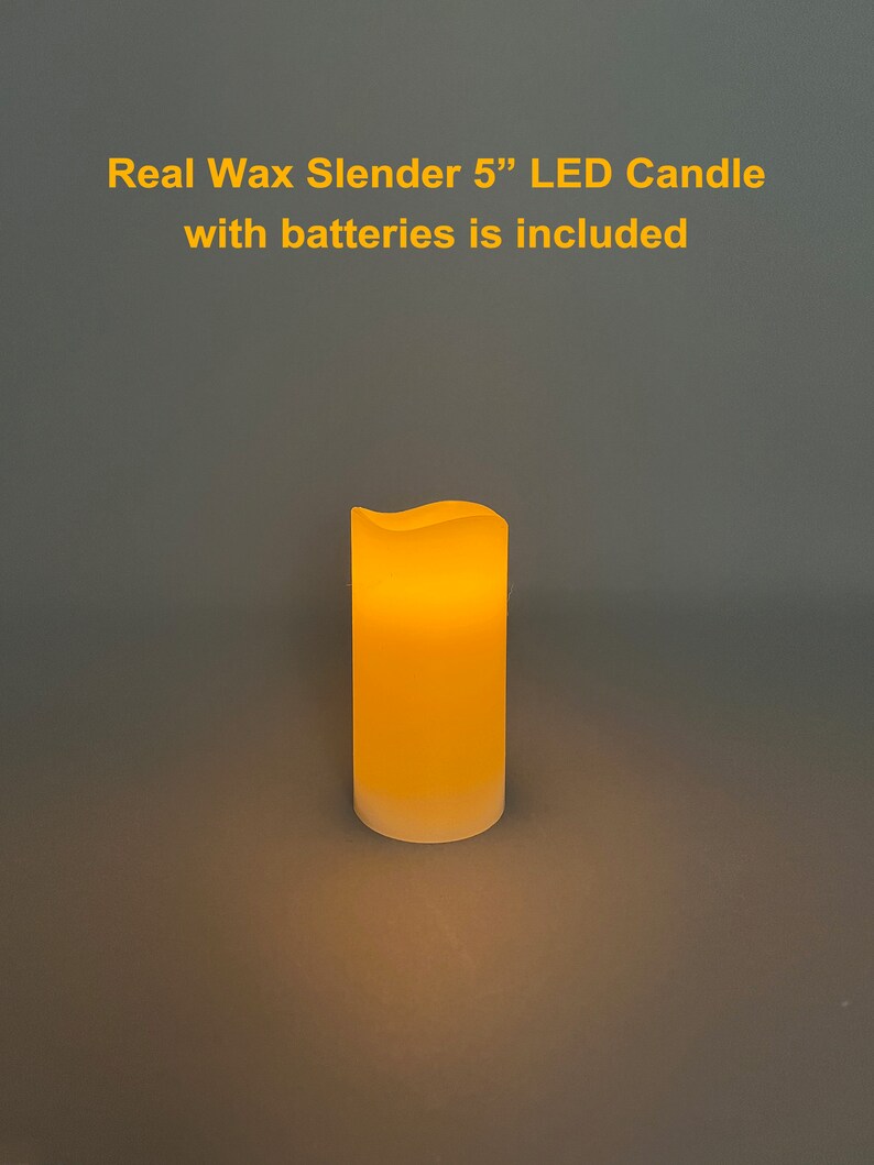 May include: A slender, real wax LED candle, approximately 5 inches tall, with a softly glowing amber light. The candle has a slightly wavy top edge and a frosted base. Text on the image reads: "Real Wax Slender 5" LED Candle with batteries is included."