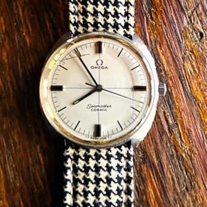 May include: A vintage Omega Seamaster Cosmic watch with a silver-toned case and a white dial. The watch features black hands and markers, and a black and white houndstooth patterned watch band. The watch is displayed on a wooden surface.