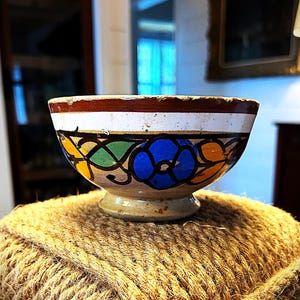 May include: A small, hand-painted ceramic bowl with a white base and a brown rim. The bowl features a floral design with blue, green, yellow, and orange accents.