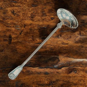 May include: A silver-toned metal ladle featuring a long, slender handle with a ribbed design and a decorative shell motif at the base. The bowl of the ladle is oval-shaped and polished to a reflective finish, showcasing a classic, elongated silhouette.
