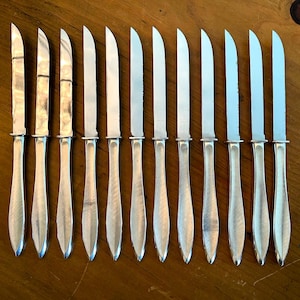 May include: A set of twelve silver-colored butter knives arranged in a row on a wooden surface. Each knife has a shiny, curved blade and a long, tapered handle. The knives are evenly spaced, creating a symmetrical pattern.