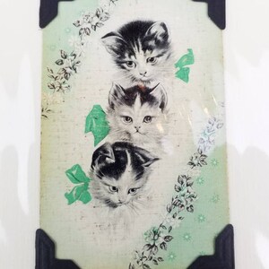 May include: Vintage print featuring three adorable kittens with black and white fur, set against a light green background with floral accents and green bows. The print is framed with black corner protectors, adding a touch of nostalgia.