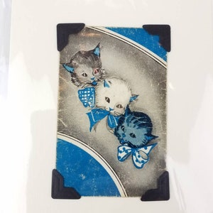 Puede incluir: Impresi&oacute;n vintage con tres gatitos de dibujos animados con lazos y detalles azules. Los gatitos tienen pelaje gris, blanco y azul. La impresi&oacute;n est&aacute; sobre un fondo gris y azul y se sujeta con esquinas negras.